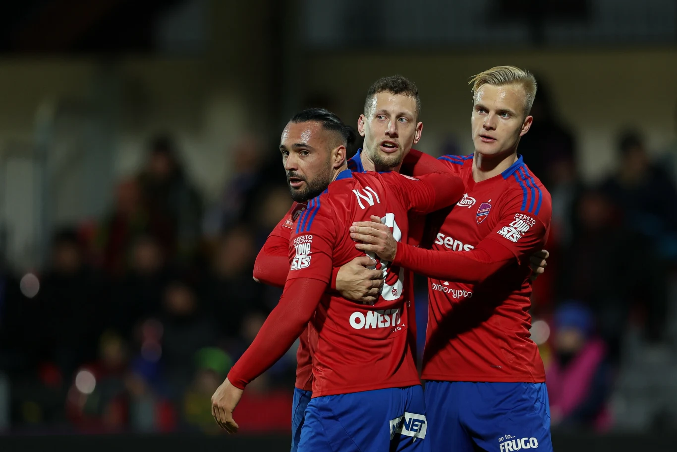 Zawodnicy Rakowa Częstochowa Trzech piłkarzy drużyny w czerwonych koszulkach z logo sponsorów obejmuje się i celebruje razem na tle rozmytej publiczności na stadionie.