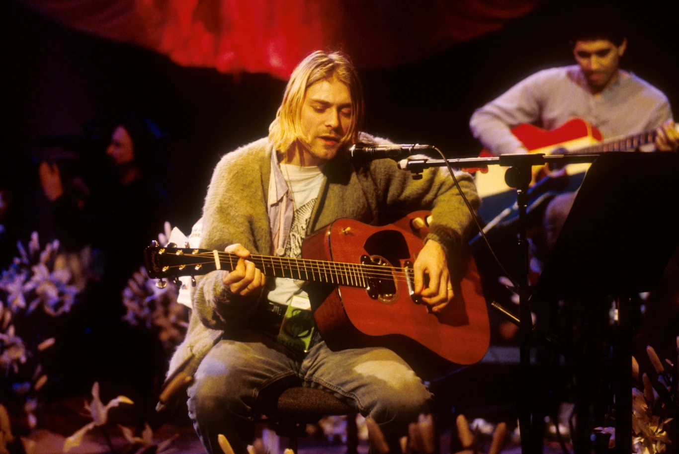 Kurt Cobain podczas koncertu Nirvany "MTV Unplugged" Muzyk z długimi blond włosami gra na gitarze akustycznej, ubrany w szary sweter i dżinsy, siedzący na scenie oświetlonej ciepłym światłem, w tle inny gitarzysta i dekoracje w kształcie kwiatów.