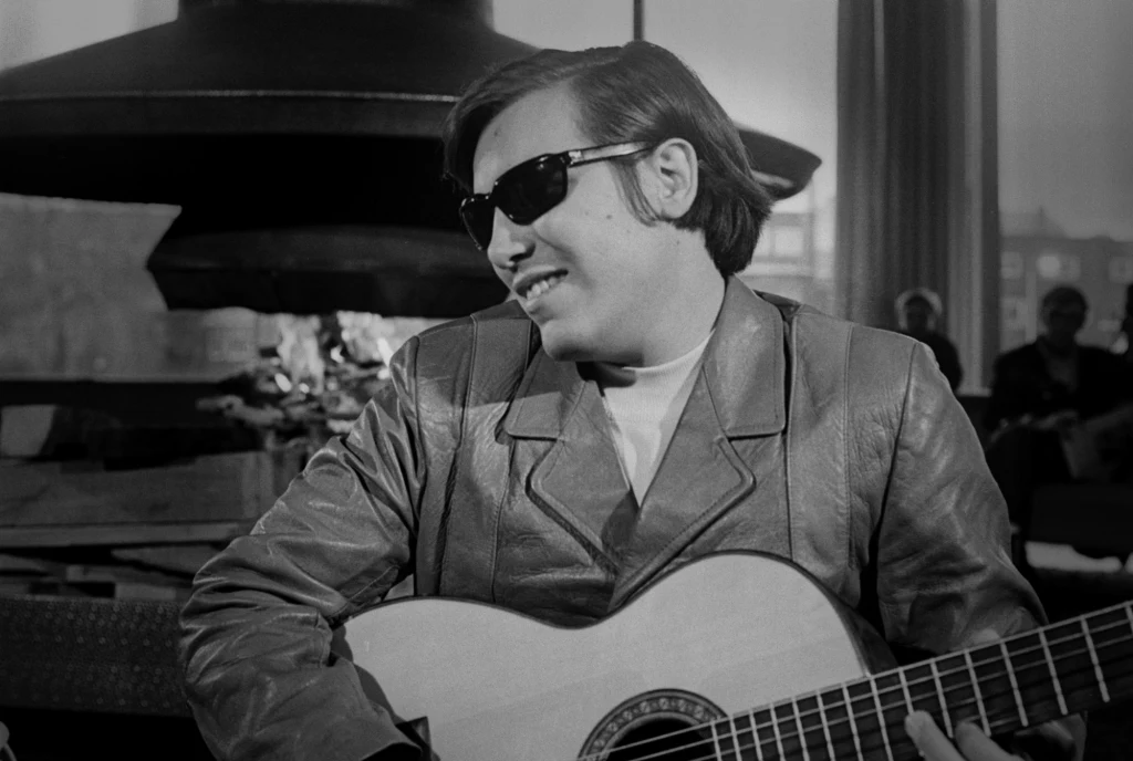 Mężczyzna w okularach przeciwsłonecznych grający na gitarze, ubrany w jasną skórzaną kurtkę, siedzi w jasnym pomieszczeniu z widocznym kominkiem w tle.