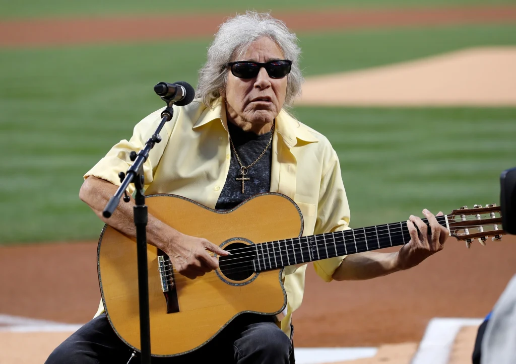 Starszy mężczyzna w okularach przeciwsłonecznych, ubrany w żółtą koszulę, gra na gitarze akustycznej i śpiewa do mikrofonu podczas występu na stadionie.