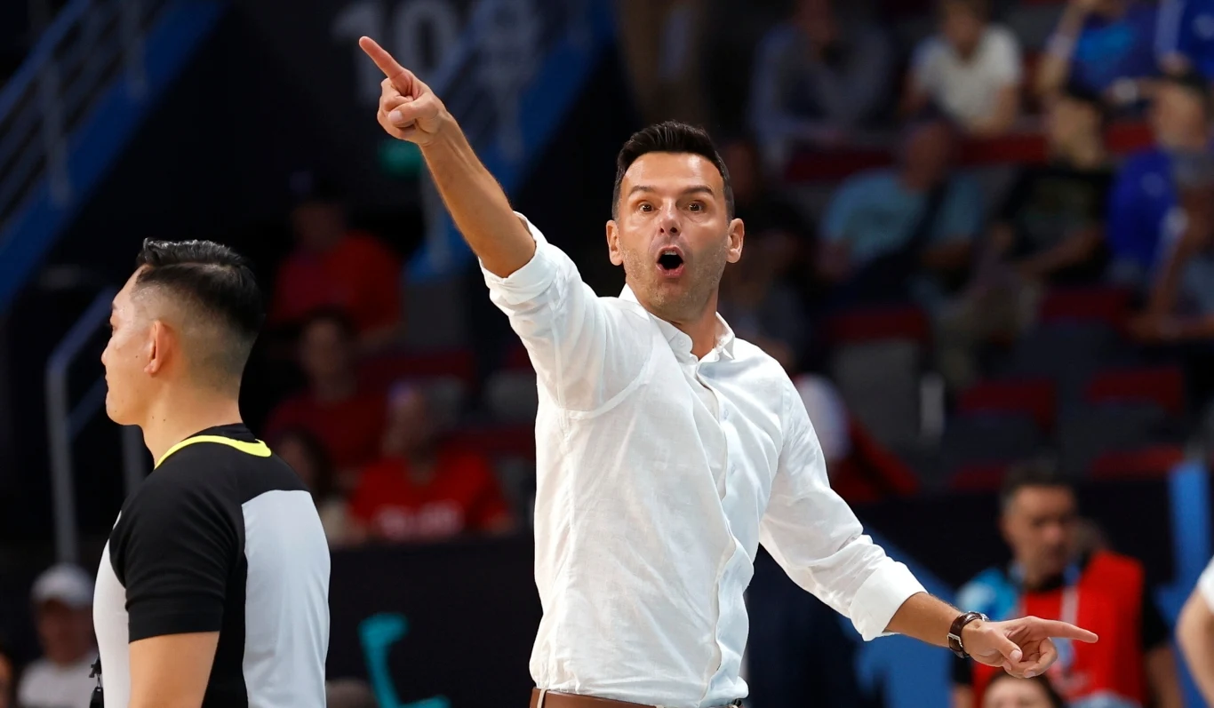 Trener energicznie gestykuluje ręką i wskazuje kierunek, na pierwszym planie widoczny jest również sędzia sportowy, a w tle rozmyta publiczność na trybunach.
