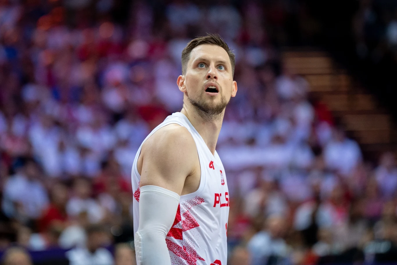 Koszykarz w białym stroju z czerwonymi elementami i napisem Polska koncentruje się na boisku sportowym, a w tle widoczna jest rozmyta publiczność na trybunach.