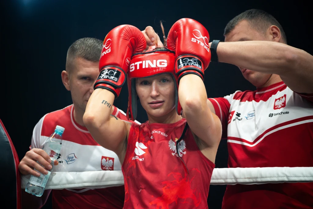 Bokserka w czerwonym stroju z orłem na piersi otoczona przez dwóch trenerów, zakładająca rękawice bokserskie z napisem STING, w tle widoczne biało-czerwone stroje z polskim godłem.