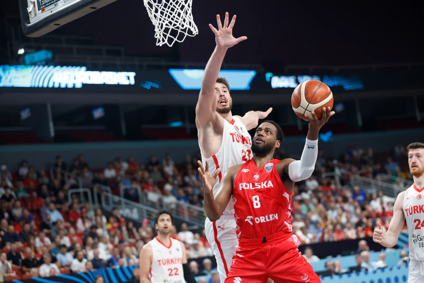 Zawodnik reprezentacji Polski próbuje zdobyć punkt spod kosza, będąc blokowanym przez gracza reprezentacji Turcji, wokół obu sportowców widoczni są inni uczestnicy meczu oraz liczna publiczność na trybunach.