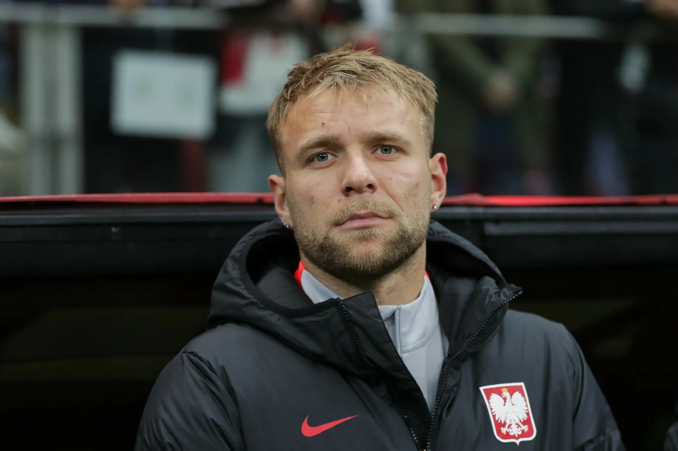 Mężczyzna w kurtce sportowej z godłem Polski, stojący w pobliżu stadionu, skoncentrowany wyraz twarzy, tło rozmyte z niewyraźnymi postaciami i elementami infrastruktury sportowej.