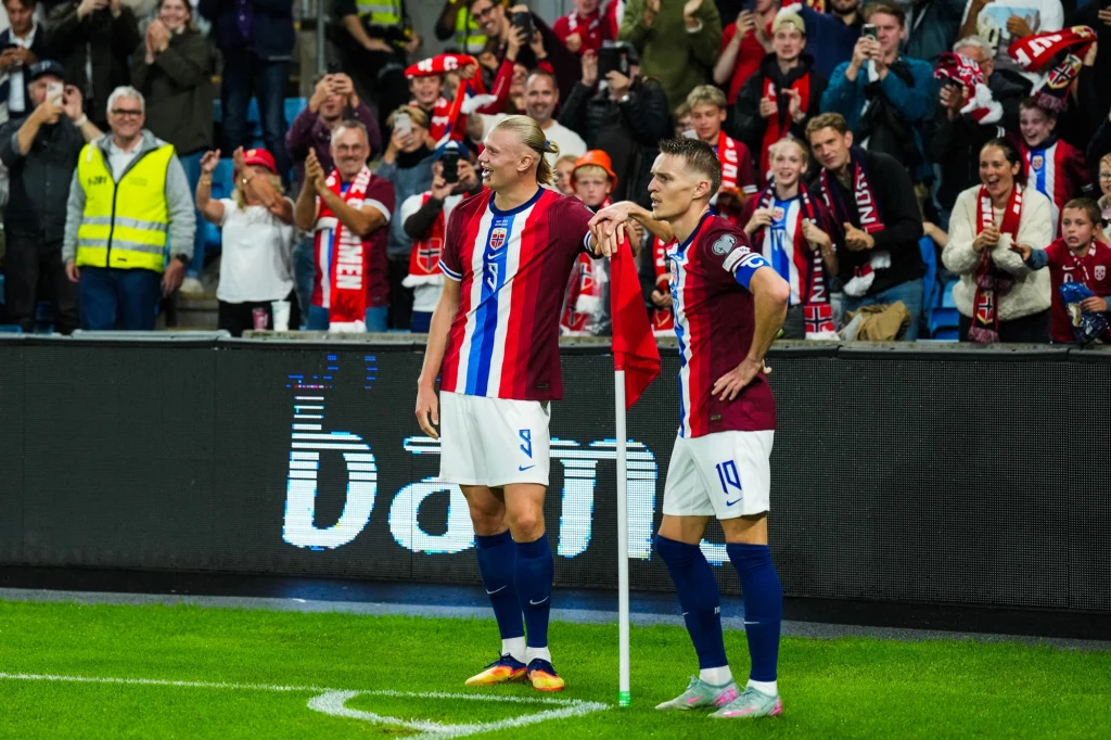 Erling Haaland i Martin Odegaard mogą wprowadzić Norwegią na mundial Dwóch piłkarzy w bordowo-niebieskich strojach świętuje przy chorągiewce narożnej na stadionie, za nimi tłum rozentuzjazmowanych kibiców ubranych w klubowe barwy.