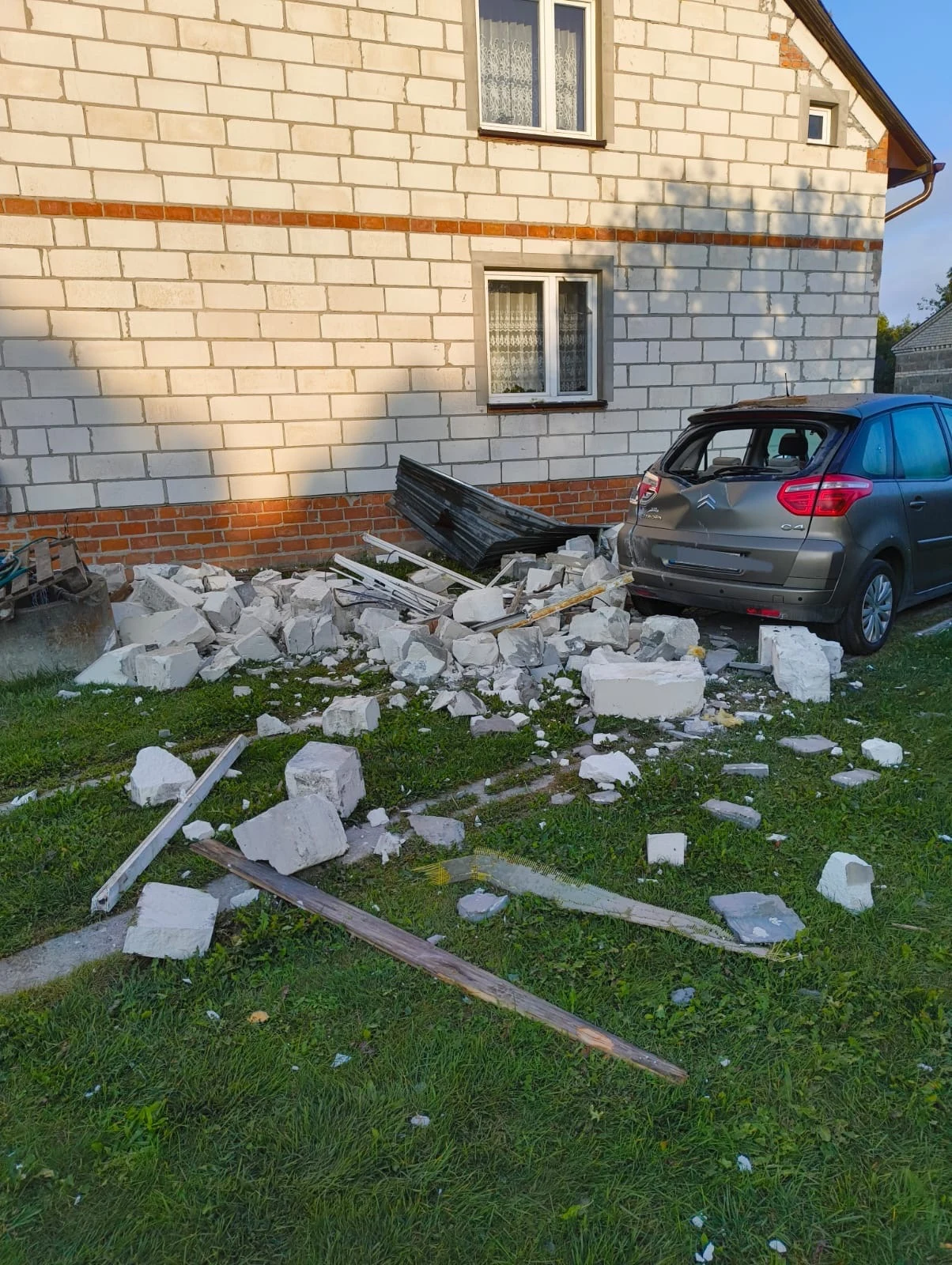 Zniszczony mur budynku oraz rozbity samochód stojący obok, wokół leżą porozrzucane fragmenty cegieł i gruzu na zielonej trawie.