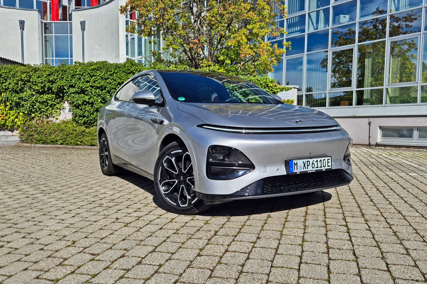 Xpenga G6 po modernizacji najłatwiej rozpoznać po pasie świetlnym, którego nie przecina już logo - trafiło ono na maskę Srebrny samochód elektryczny zaparkowany na brukowanym placu przed nowoczesnym budynkiem ze szklanymi elementami, otoczony zielenią i drzewami.