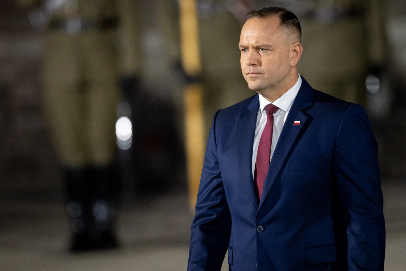 Prezydent Karol Nawrocki reaguje na naruszenie polskiej przestrzeni Mężczyzna w granatowym garniturze z czerwoną krawatą i białą koszulą, z przypiętą polską flagą na klapie, stoi na tle żołnierzy w mundurach podczas oficjalnej uroczystości.