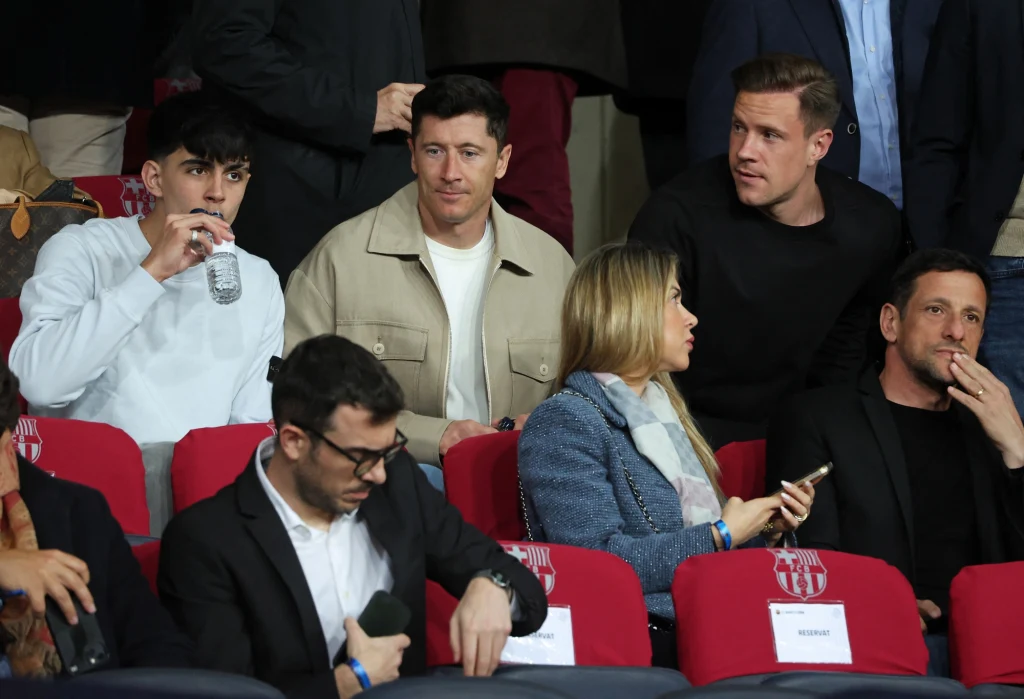 Marc Bernal, Robert Lewandowski, Marc-Andre ter Stegen Grupa kilku osób siedzących na czerwonych krzesłach na widowni, część osób rozmawia ze sobą, jedna osoba pije wodę z butelki, inni patrzą w różne kierunki, na siedzeniach widoczne są kartki z napisami rezerwującymi miejsce.