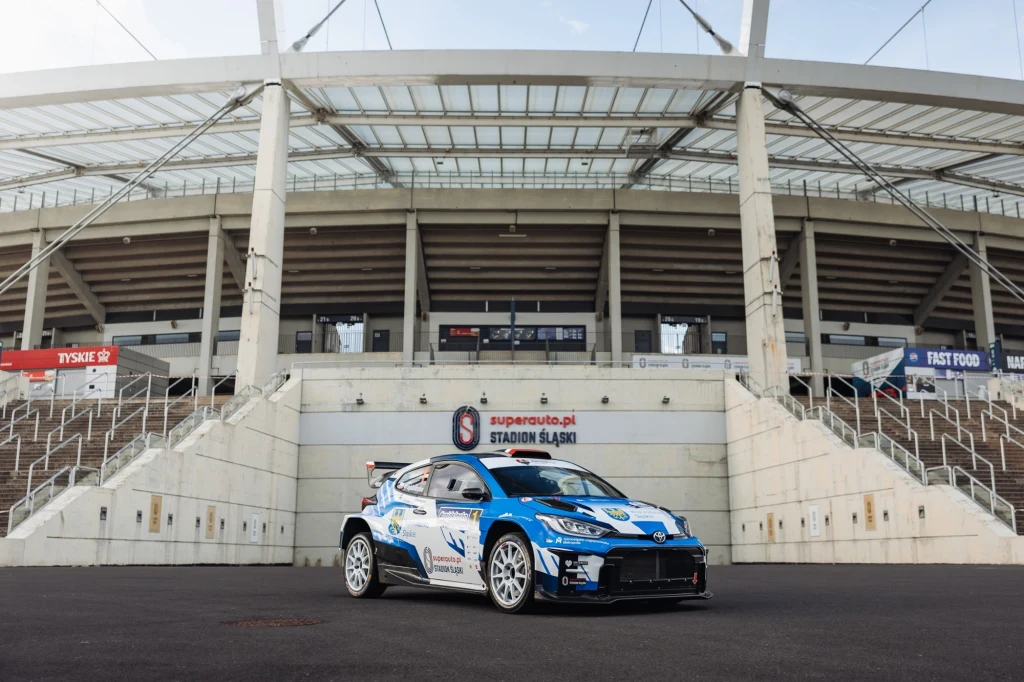 Auto rajdowe w niebiesko-białych barwach stoi na tle wejścia głównego Stadionu Śląskiego, nowoczesna konstrukcja trybun i schody prowadzące do wejścia widoczne w tle.