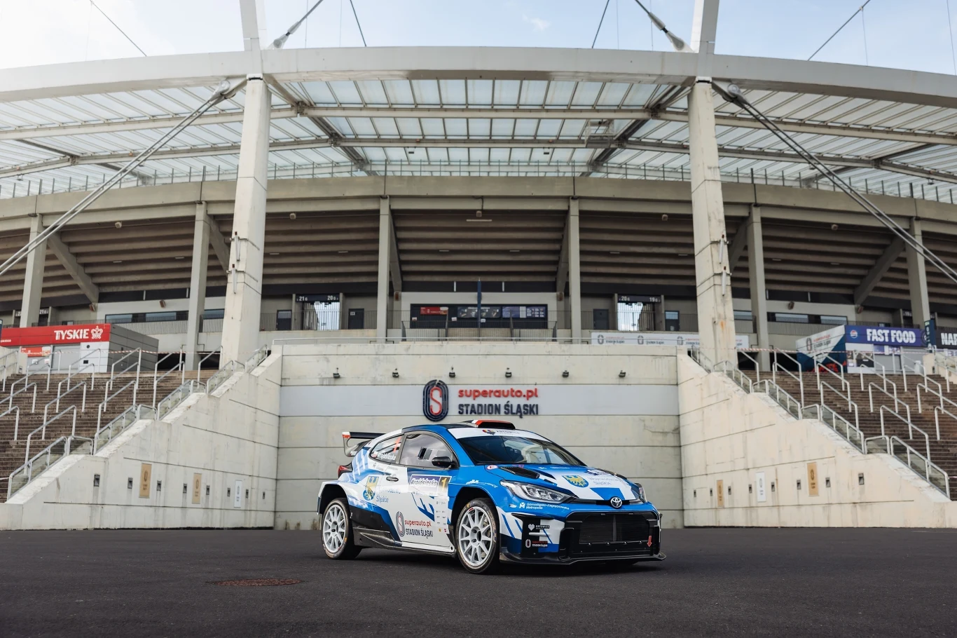 Rajd Śląska 2025 Auto rajdowe w niebiesko-białych barwach stoi na tle wejścia głównego Stadionu Śląskiego, nowoczesna konstrukcja trybun i schody prowadzące do wejścia widoczne w tle.