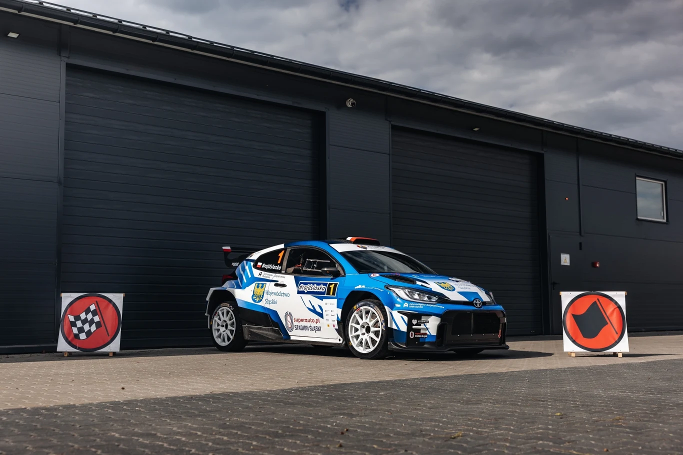 Sportowy samochód rajdowy w kolorze niebieskim z licznymi naklejkami sponsorów, ustawiony na tle nowoczesnego budynku z zamkniętymi, czarnymi bramami garażowymi, po bokach stoją znaki związane z wyścigami.