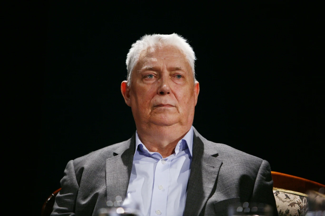 Wojciech Młynarski mógł napisać tekst hitu "Do zakochania jeden krok" Starszy mężczyzna o siwych włosach w eleganckim garniturze i jasnej koszuli siedzi na tle ciemnego, neutralnego tła, patrząc poważnie przed siebie.