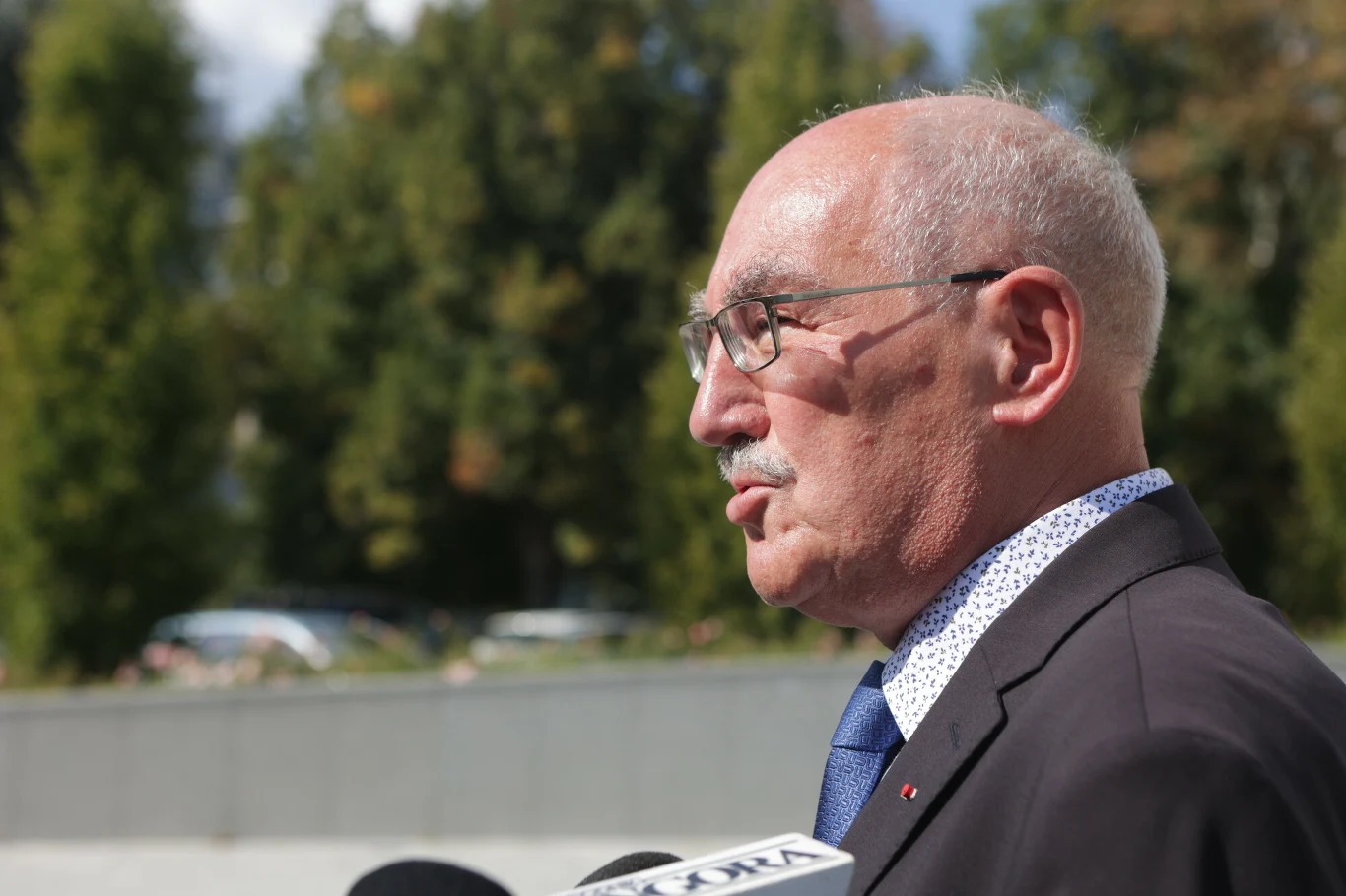 Starszy mężczyzna w garniturze i okularach, udzielający wywiadu na tle drzew i jasnego nieba, trzyma mikrofon przed sobą.