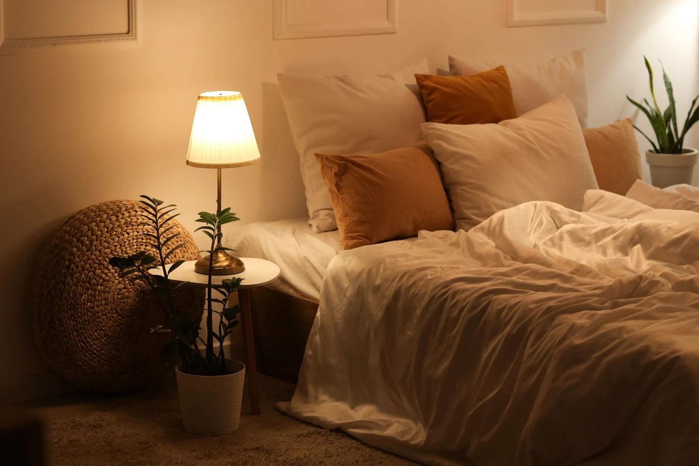 Jak ustawić łóżko w sypialni według feng shui? Łóżko z białą pościelą, brązowymi poduszkami i oświetlone lampką nocną, obok stolik z rośliną doniczkową, tworzące przytulny, ciepły klimat sypialni.