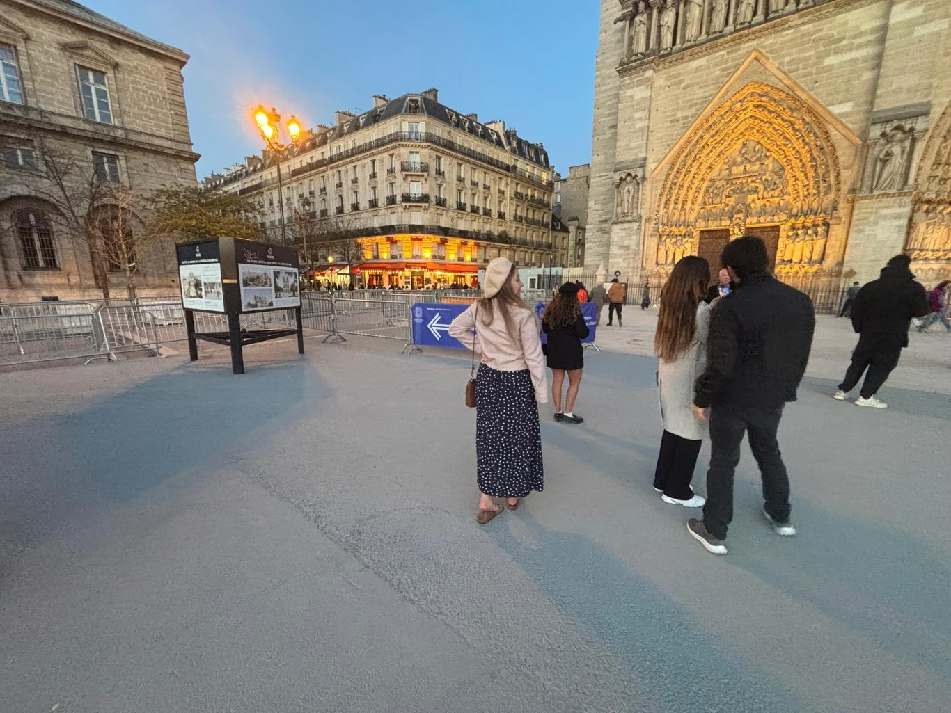 Młoda kobieta w długiej spódnicy i jasnej bluzie pozuje na placu przy katedrze Notre-Dame w Paryżu, w tle widoczne są zabytkowe budynki, podświetlona elewacja katedry oraz kilkoro przechodniów.