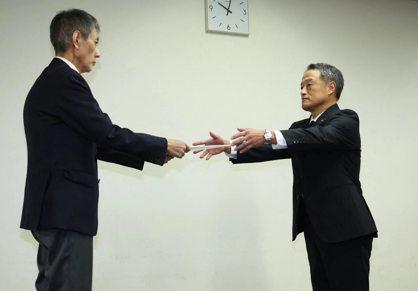 Dwóch mężczyzn w garniturach stoi naprzeciwko siebie w pomieszczeniu, jeden z nich przekazuje drugiemu dokument, nad nimi na ścianie wisi zegar.