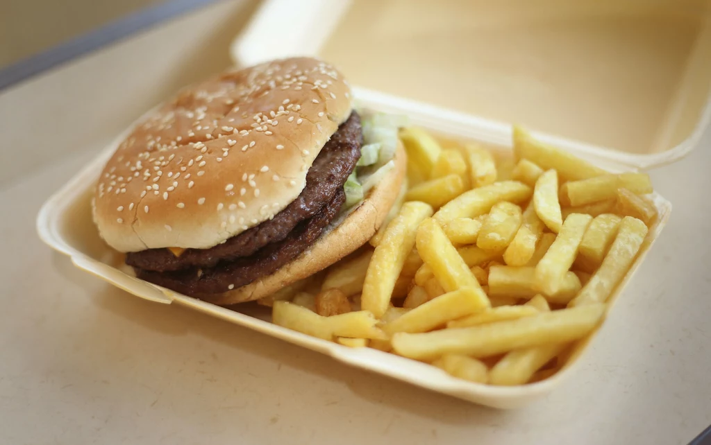 Wysokie spożycie tych produktów wiąże się ze zwiększonym ryzykiem otyłości Podwójny hamburger z bułką sezamową oraz porcją frytek w plastikowym opakowaniu typu fast food.