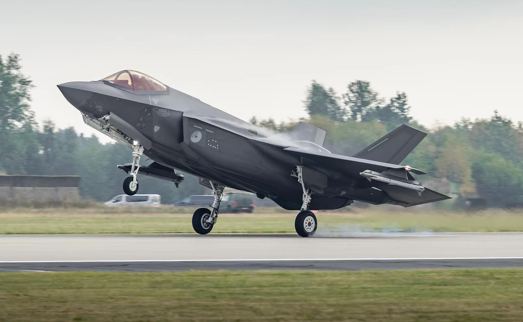 Holenderski F-35 Myśliwiec wielozadaniowy startujący lub lądujący na pasie lotniska, w tle widoczne drzewa i niewyraźne budynki.