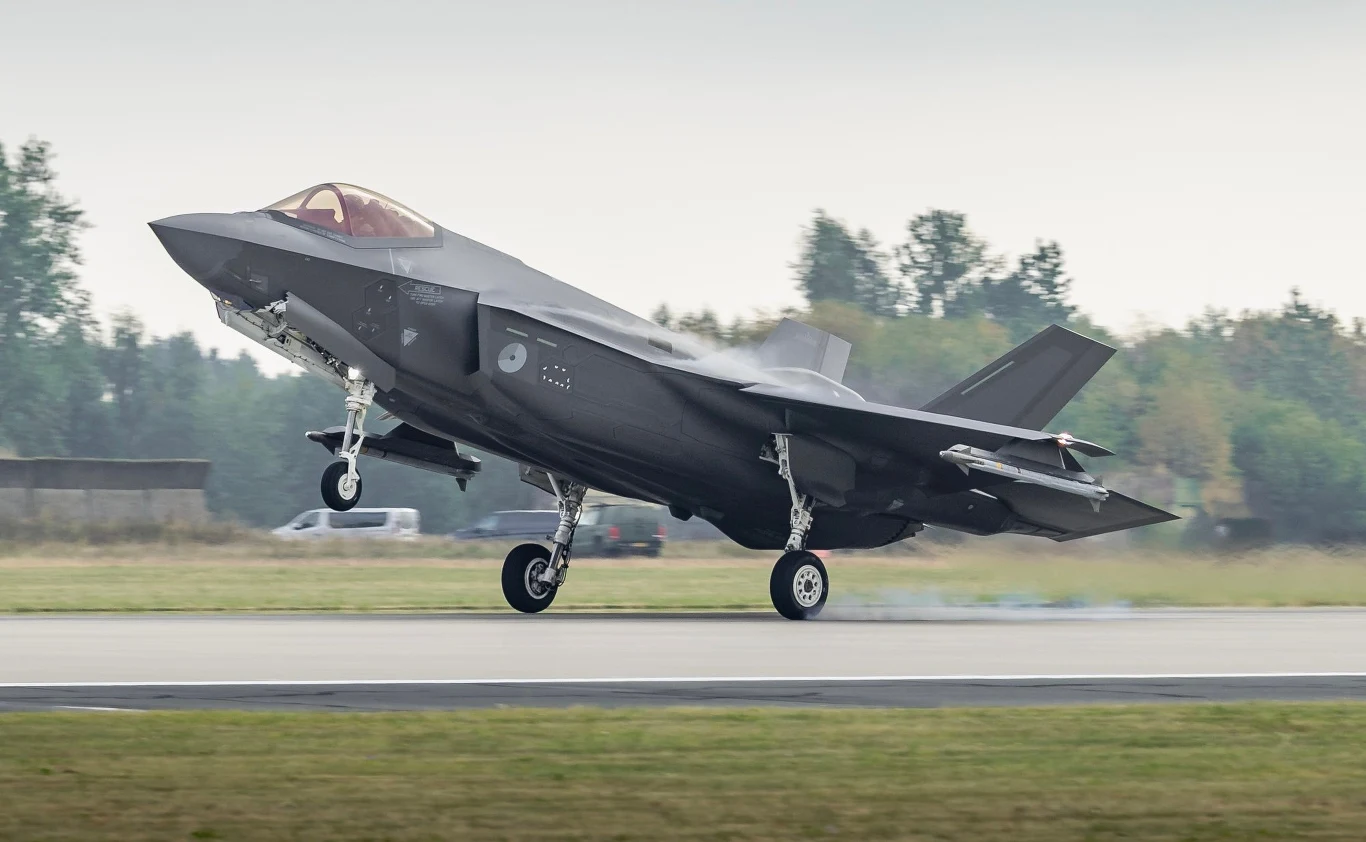 Holenderski F-35 Myśliwiec wielozadaniowy startujący lub lądujący na pasie lotniska, w tle widoczne drzewa i niewyraźne budynki.