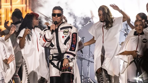 Wokalista w futurystycznym stroju śpiewa na scenie, otoczony przez tancerki ubrane na biało w peleryny i srebrne kozaki, dynamiczne światła i dym sceniczny tworzą energetyczną atmosferę występu muzycznego.