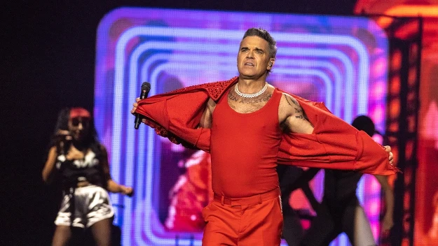 Mężczyzna w czerwonym stroju scenicznym trzyma mikrofon na tle kolorowych, geometrycznych świateł, w tle tancerze i dynamiczna atmosfera koncertu.