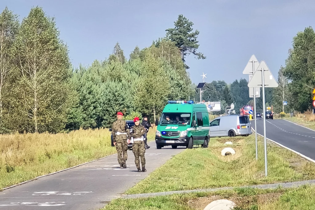 Dwóch żołnierzy w mundurach chodzi ścieżką rowerową przy drodze, obok stoi policyjny radiowóz i inne pojazdy służbowe, otoczenie stanowi zieleniec i drzewa, atmosfera spokojna.