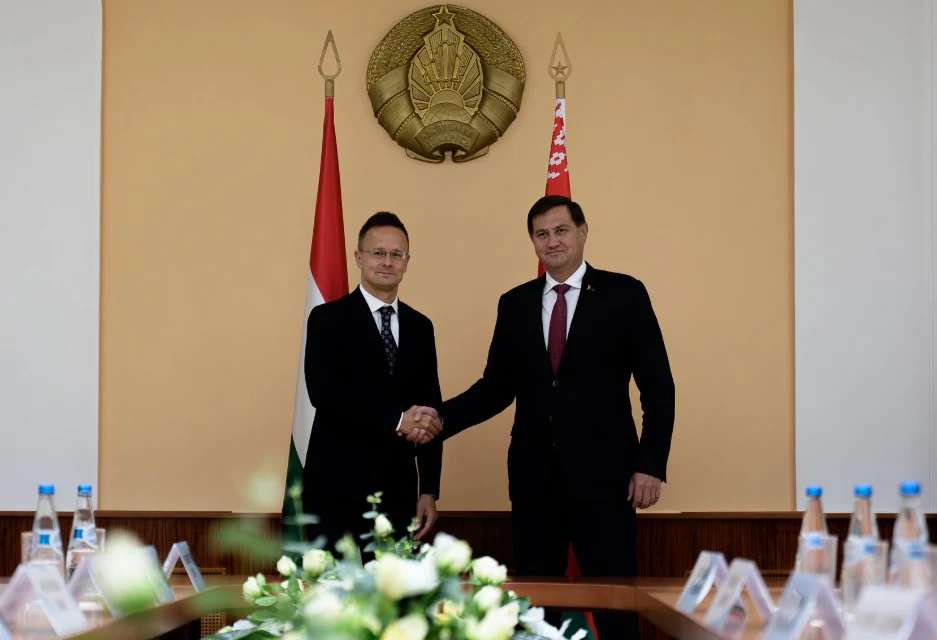 Ministrowie spraw zagranicznych Węgier i Białorusi spotkali się w Mińsku Dwóch mężczyzn w garniturach pozuje do zdjęcia podczas uścisku dłoni w sali konferencyjnej, w tle widoczne flagi państwowe oraz godło na ścianie, na stole stoją plastikowe butelki z wodą i plakietki.