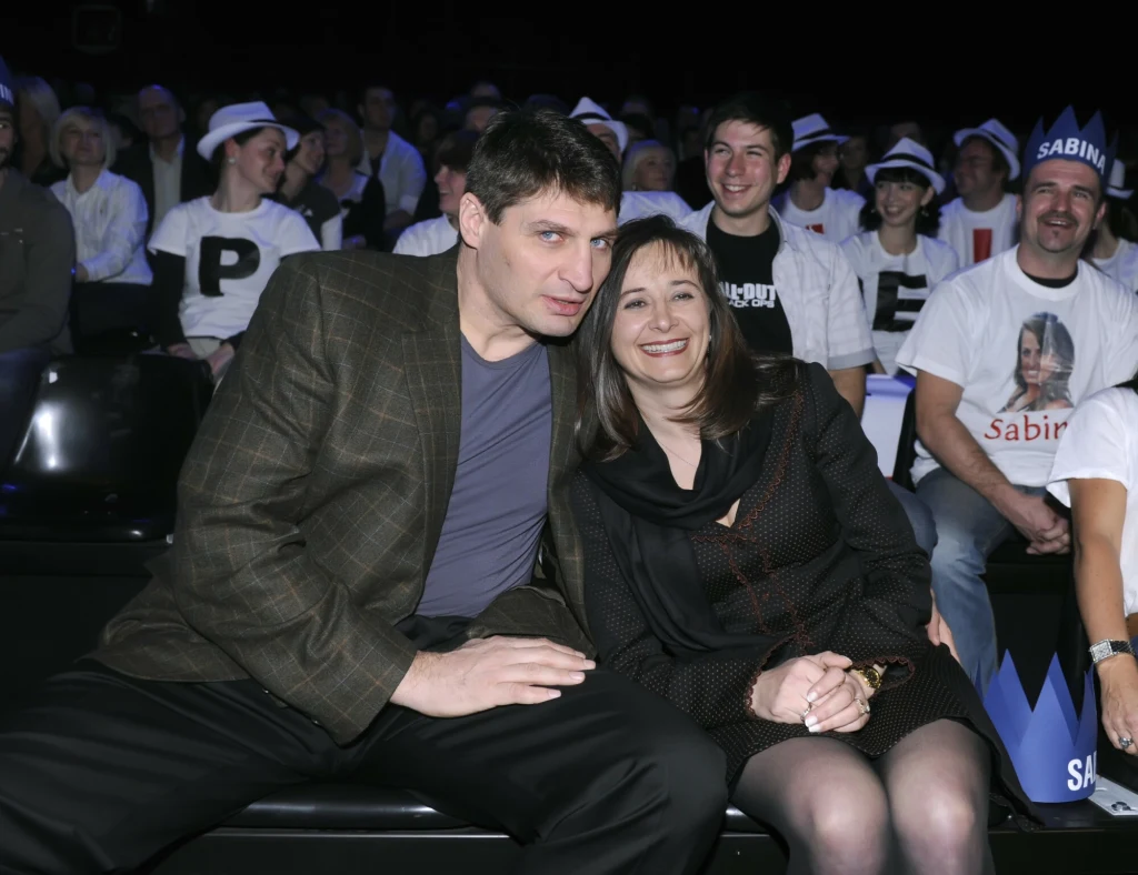 Para siedzi razem na widowni, uśmiechnięci, otoczeni grupą kibiców ubranych w białe koszulki oraz charakterystyczne kapelusze, w tle widać tabliczki i elementy sugerujące doping dla uczestnika wydarzenia.