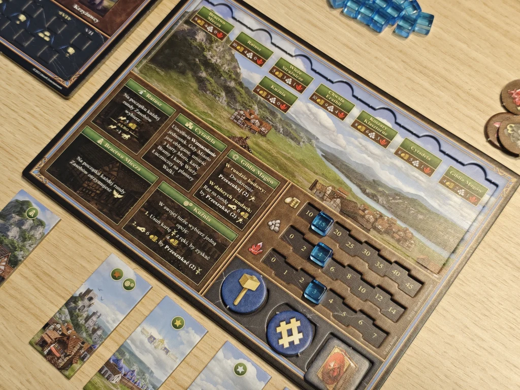 Elementy planszy do gry planszowej z widocznymi kartami, znacznikami oraz planszą z torami postępu i instrukcjami. Na planszy widoczne są malownicze krajobrazy gór, kilka ilustracji oraz elementy gry logicznej.