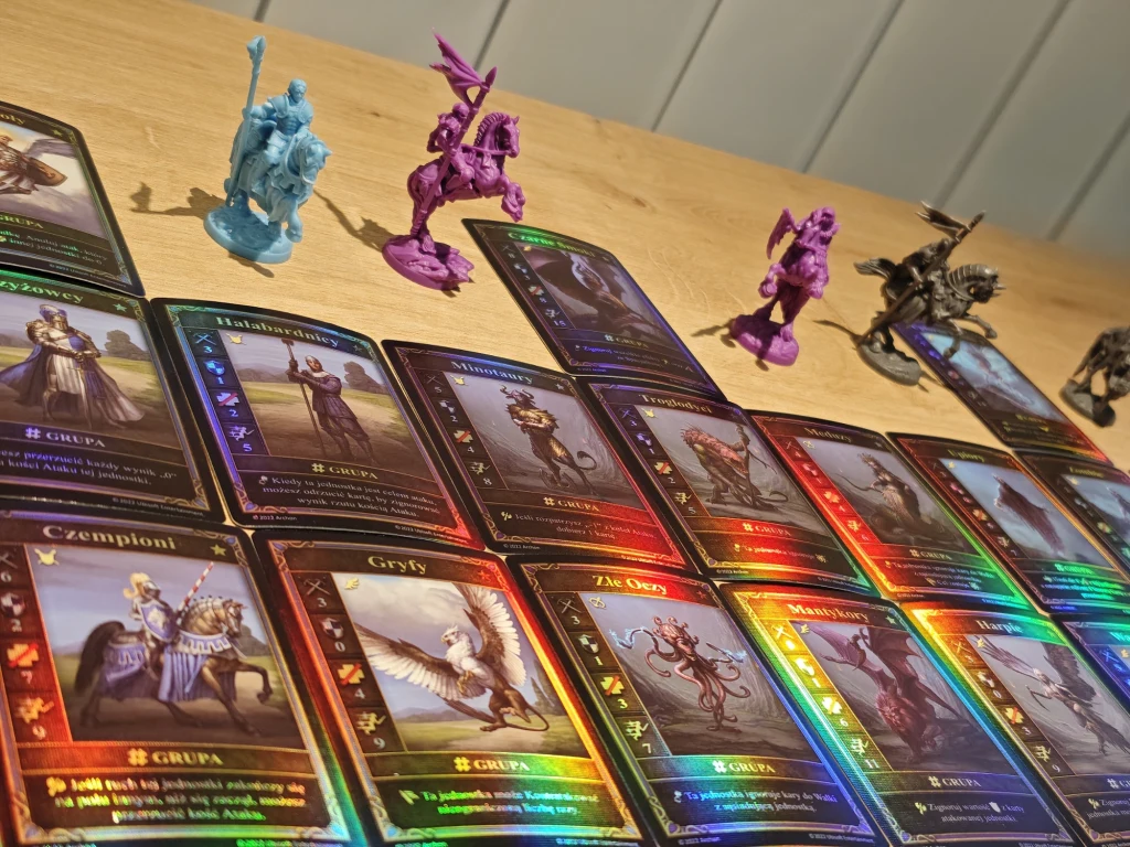 Kolorowe karty do gry strategicznej rozmieszczone na drewnianym blacie, w tle cztery plastikowe figurki fantastycznych postaci i stworzeń w różnych kolorach; światło podkreśla holograficzne elementy kart.