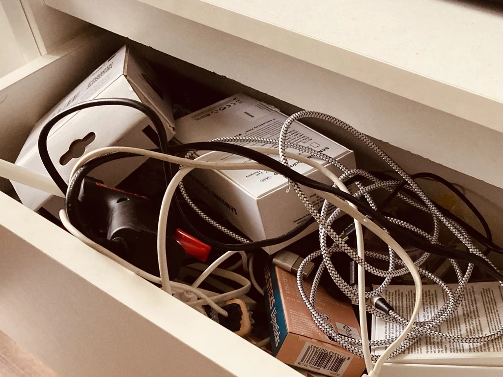 Szuflada wypełniona splątanymi kablami, ładowarkami i różnego rodzaju pudełkami elektronicznymi, tworząca wrażenie nieładu i chaosu.