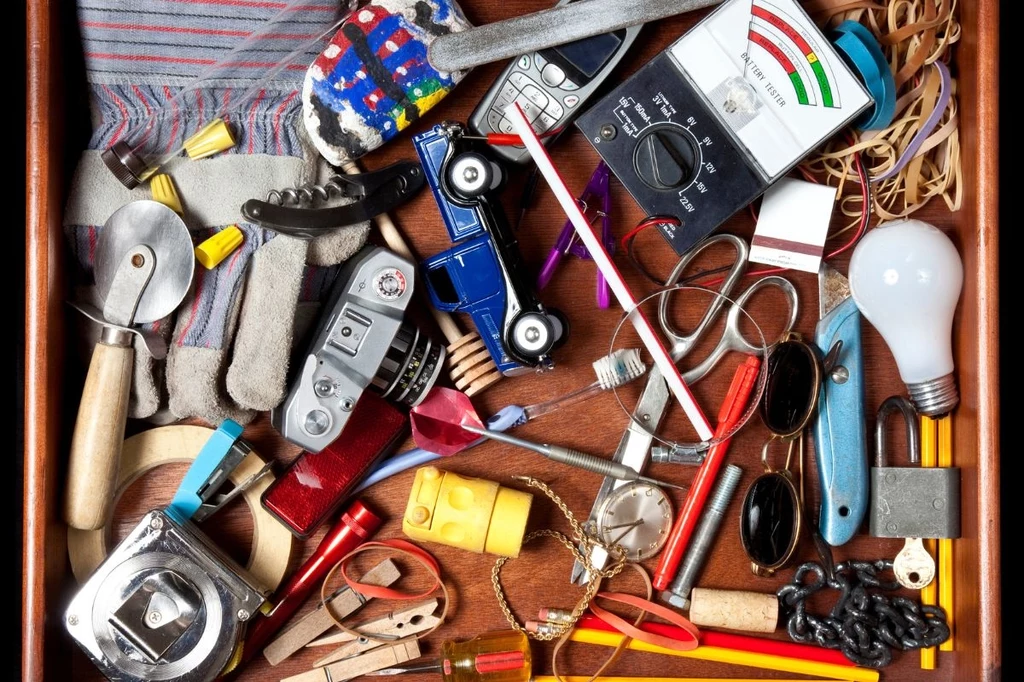 Różnorodne przedmioty codziennego użytku rozrzucone w szufladzie, w tym stara kamera, otwieracz, samochodzik, okulary przeciwsłoneczne, żarówka, taśma miernicza, telefon komórkowy, karty do gry i narzędzia.