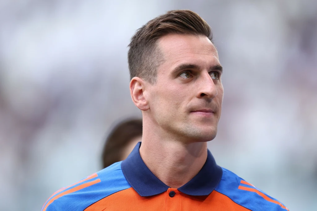 Arkadiusz Milik Mężczyzna w sportowej oficjalnej koszulce, patrzący lekko w bok z neutralnym wyrazem twarzy, tło rozmyte i jasne, skupienie na twarzy i górnej części ciała.