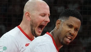 Dwóch siatkarzy w białych koszulkach z czerwonymi elementami, jeden z nich z otwartymi ustami w geście emocjonalnego okrzyku, a drugi z intensywnym wyrazem twarzy, w tle rozmyte sylwetki i siatka.