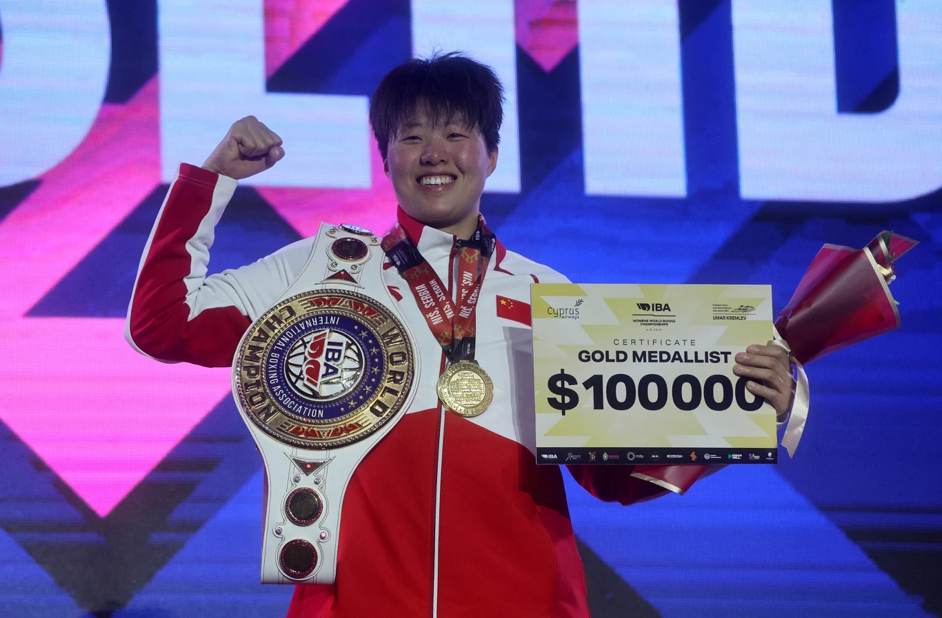 Chinka Yilian Zhan została w marcu mistrzynią świata IBA. Teraz w to samo celuje w Liverpoolu Sportowiec w czerwono-białym stroju trzyma pas mistrzowski, złoty medal, bukiet kwiatów i certyfikat informujący o nagrodzie za zwycięstwo w wysokości 100000 dolarów. Osoba uśmiecha się i unosi triumfalnie pięść.