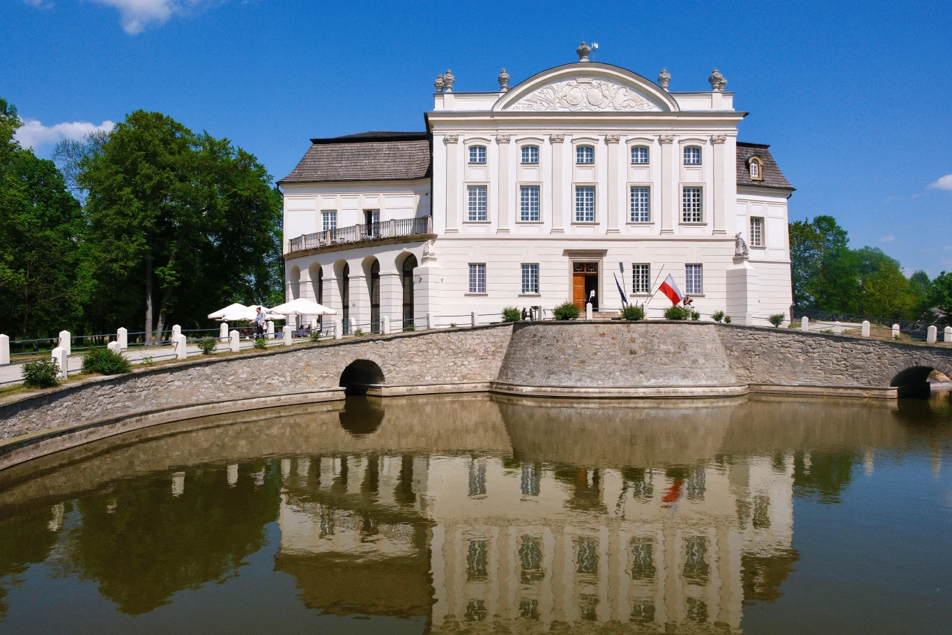 Biała, klasycystyczna rezydencja z licznymi oknami i dekoracjami architektonicznymi odbija się w spokojnej wodzie fosy, otoczona zielenią drzew oraz modnym tarasem z parasolami przy bocznym wejściu.