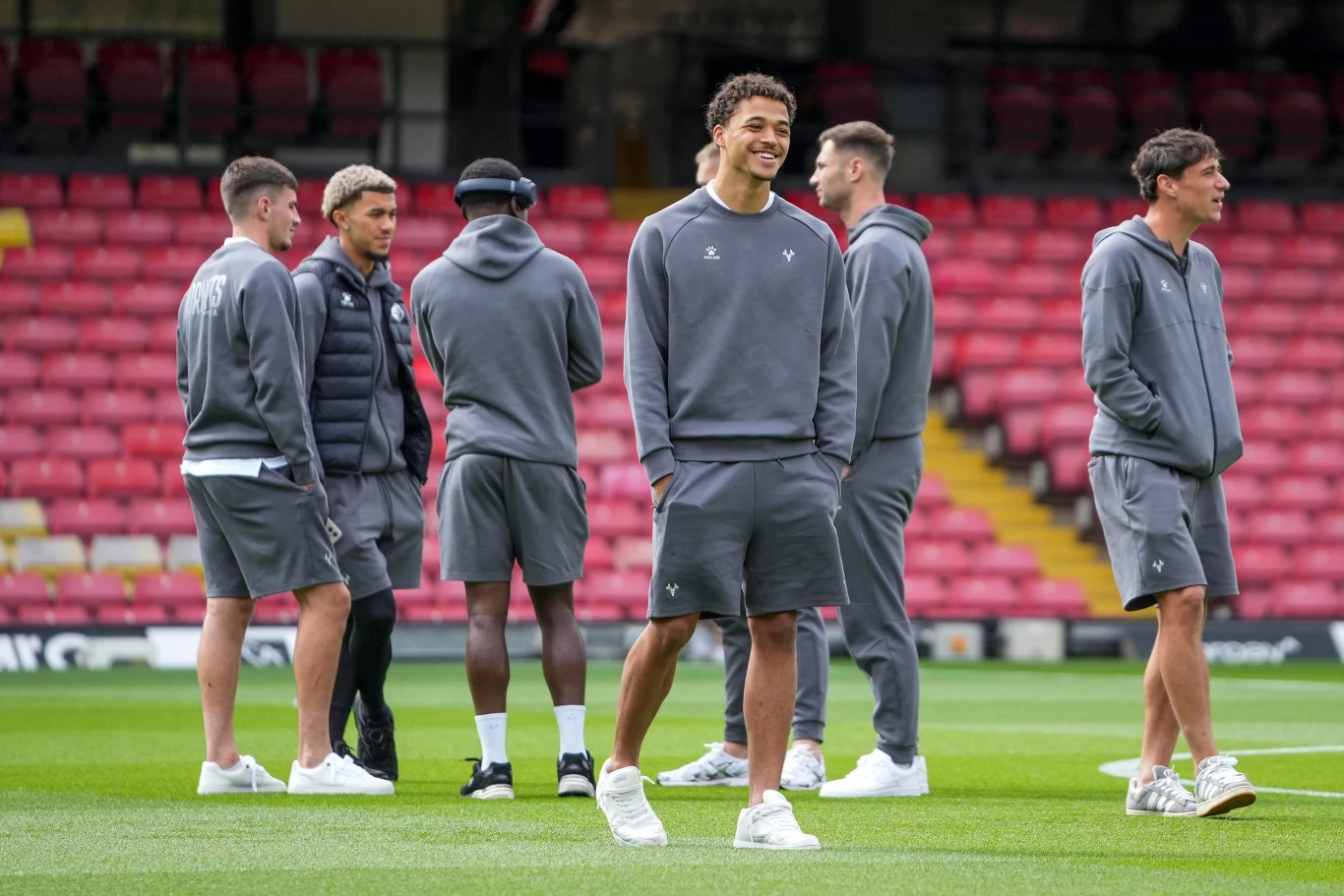 Piłkarze Watford szkoleni są zgodnie z określonymi zasadami Grupa młodych mężczyzn ubranych w sportowe dresy stoi na murawie stadionu, prowadząc luźną rozmowę wśród pustych czerwonych trybun, jeden z nich uśmiecha się szeroko.