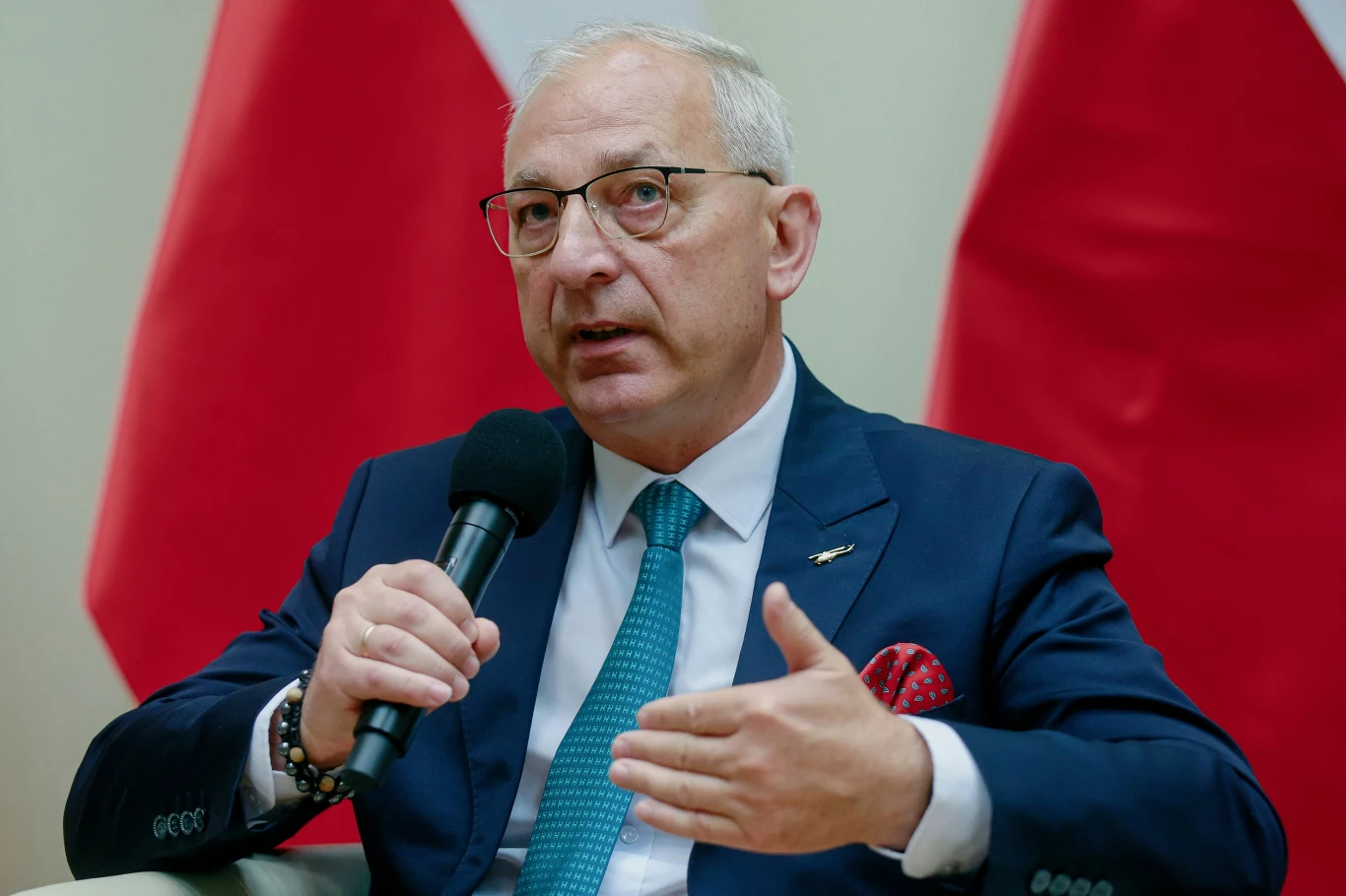 Starszy mężczyzna w okularach i granatowym garniturze siedzi i przemawia do mikrofonu, w tle widoczne są biało-czerwone flagi.