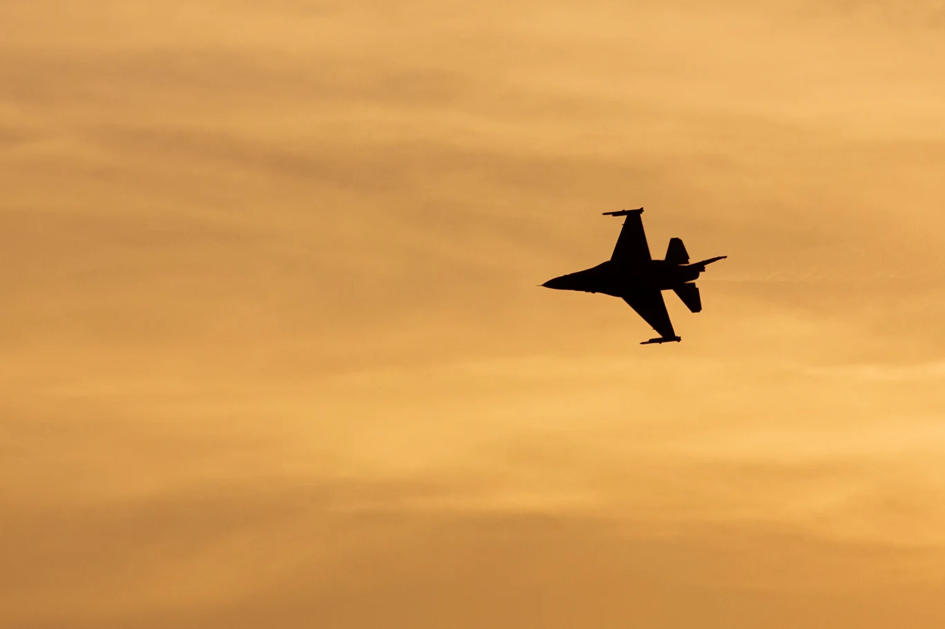 Jeden z polskich F-16 Sylwetka myśliwca odrzutowego lecącego na tle złotego, zachmurzonego nieba podczas zachodu słońca.