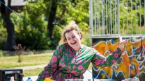 Młoda kobieta w kolorowej sukience z uśmiechem pokazuje obraźliwy gest środkowego palca stojąc przy stole na zewnątrz, wokół znajdują się rozmaite produkty spożywcze i akcesoria na tle ogrodzenia z graffiti.