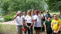 Grupa dzieci i dorosłych stoi na terenie zielonym w parku podczas wspólnej akcji sadzenia roślin, na pierwszym planie widoczne narzędzia ogrodnicze oraz wykopany dół pod sadzonkę.