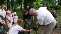 Grupa osób sadzi drzewo w parku, dzieci i dorośli współpracują, otaczają młode drzewko, część klęczy i trzyma sadzonkę, jeden z mężczyzn używa łopaty przy wsparciu pozostałych uczestników akcji.