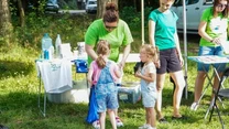 Dwie dziewczynki uczestniczą w aktywności plenerowej prowadzonej przez kobietę w zielonej koszulce, wokół znajdują się plansze edukacyjne oraz stolik z materiałami, całość rozgrywa się w parku lub lesie.