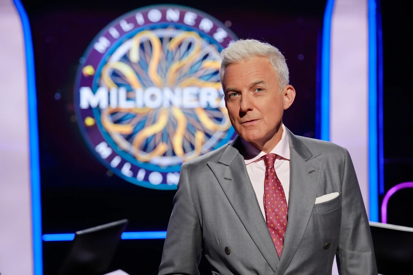 Hubert Urbański prowadzi "Milionerów" w Polsacie Mężczyzna o siwych włosach w eleganckim garniturze i różowym krawacie stoi na tle charakterystycznego logotypu programu Milionerzy z jasnym oświetleniem sceny i fioletowymi akcentami w tle.