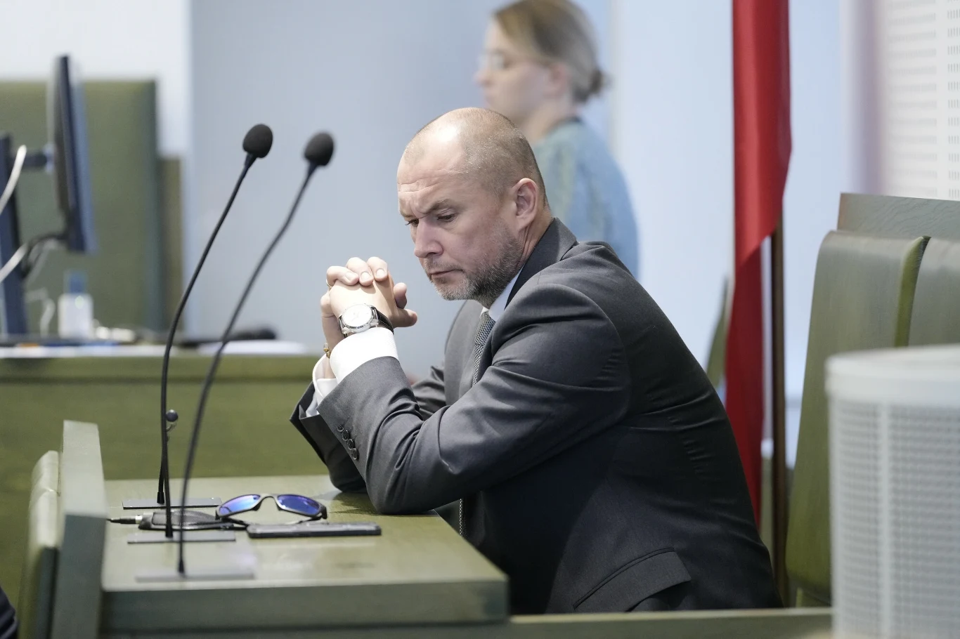 Jakub Iwaniec Mężczyzna w garniturze siedzi przy stole w sali konferencyjnej lub sądowej, opierając łokcie na blacie i składając dłonie w zamyśleniu, na stole leżą okulary przeciwsłoneczne; w tle widoczna niewyraźnie kobieta na stojąco oraz mikrofony.