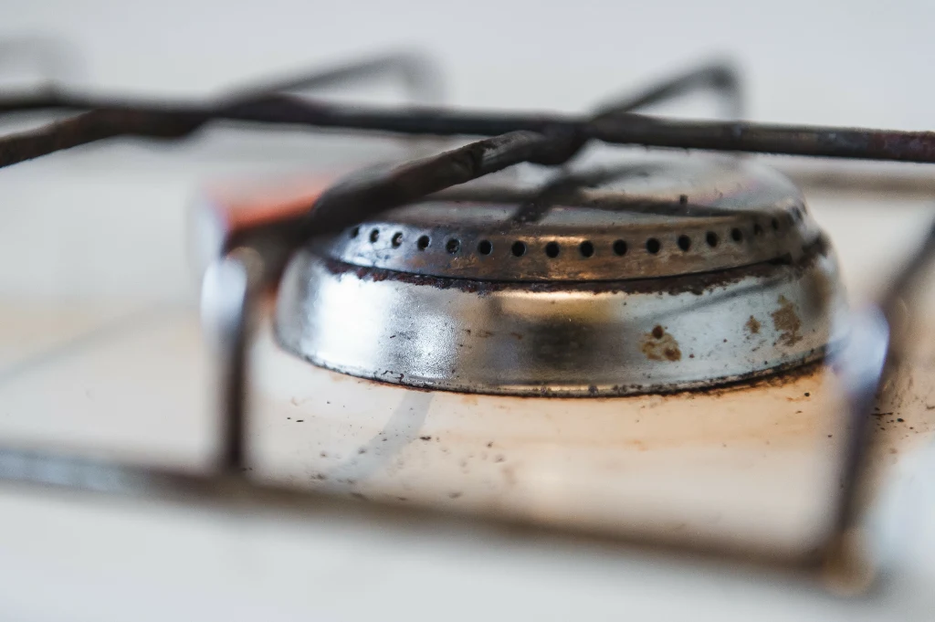 Metalowy palnik kuchenki gazowej z widoczną kratką ochronną, lekko zabrudzona powierzchnia oraz rozmyte tło wskazujące na domowe warunki.