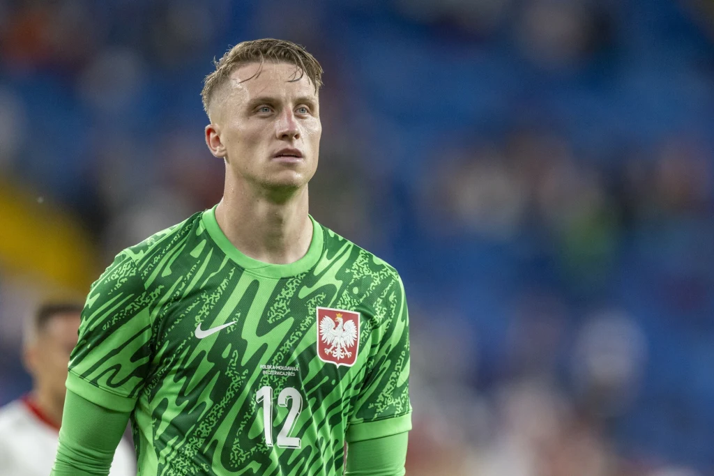 Marcin Bułka Piłkarz w zielonym stroju bramkarza z numerem 12 oraz godłem Polski na piersi, stojący na tle rozmytej publiczności w stadionie.