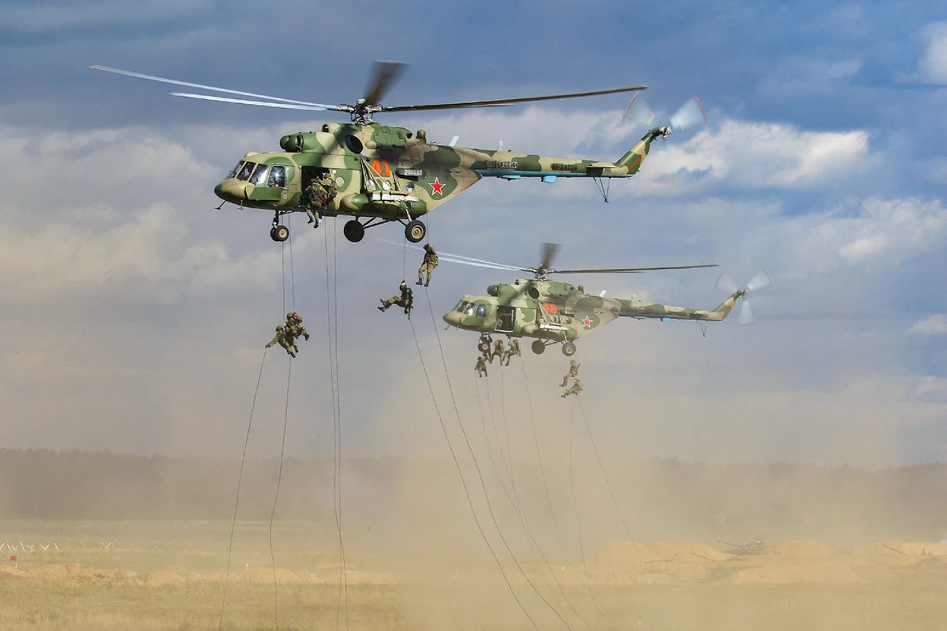 Rosyjsko-białoruskie manewry Zapad w 2021 roku Dwa wojskowe śmigłowce unoszą się nad pustynnym terenem, z których żołnierze zjeżdżają na linach w trakcie ćwiczeń, gęsty pył unosi się spod śmigłowców, a w tle widać rozległy krajobraz i jasne niebo.